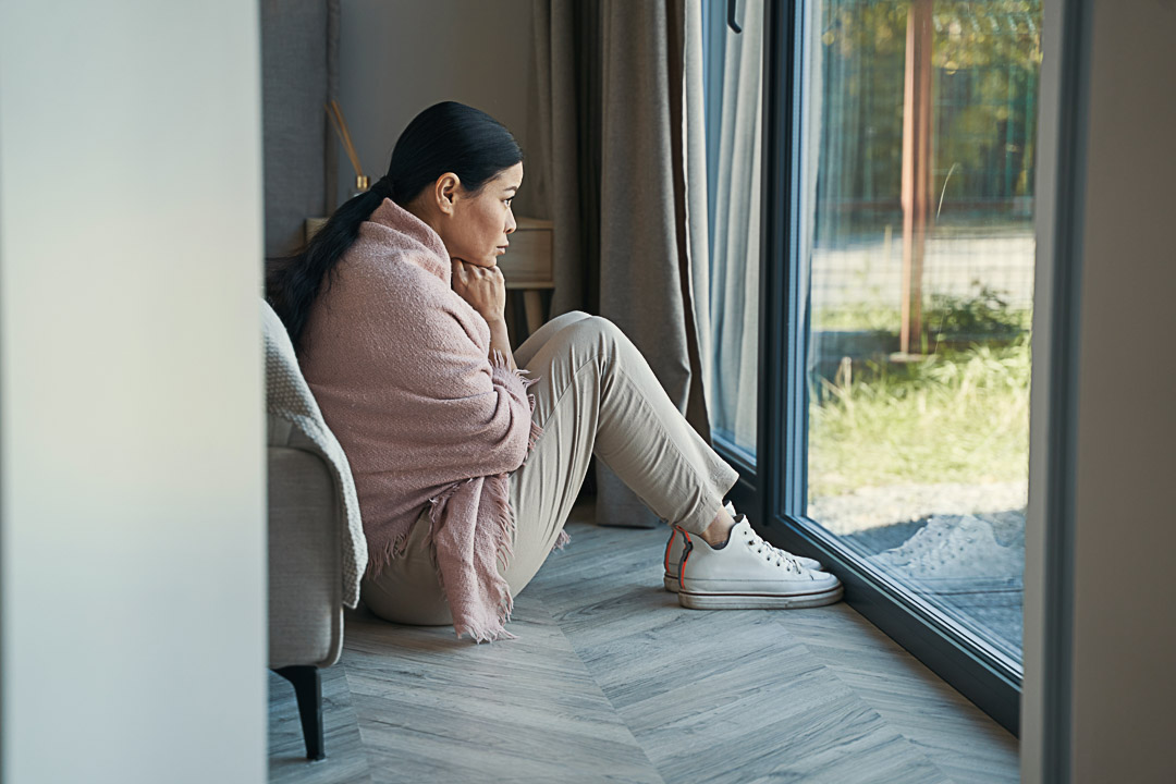 Sad mature female is sitting on floor in room and looking through window during day at home