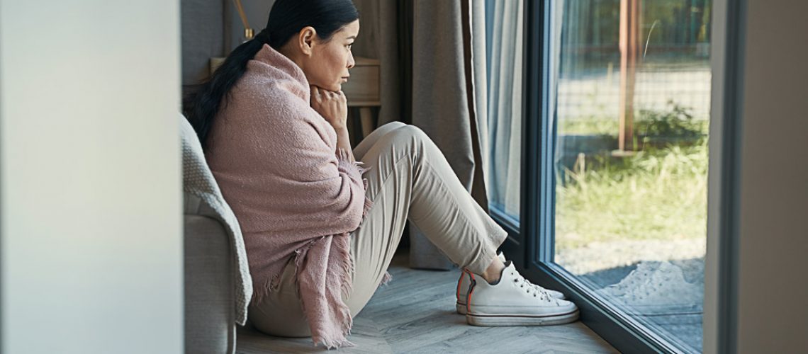 Sad mature female is sitting on floor in room and looking through window during day at home